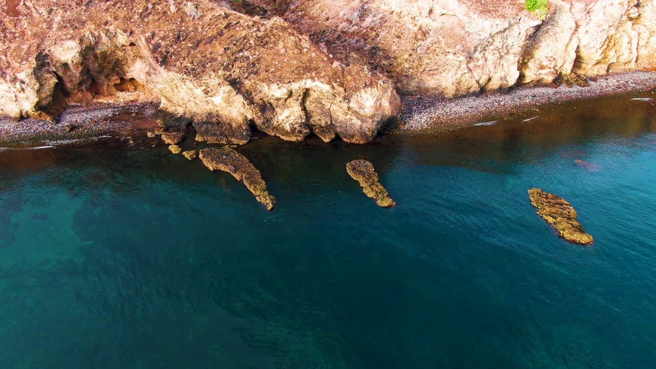 Rock cliff edge, narrow shore, and clear ocean with small ripples.