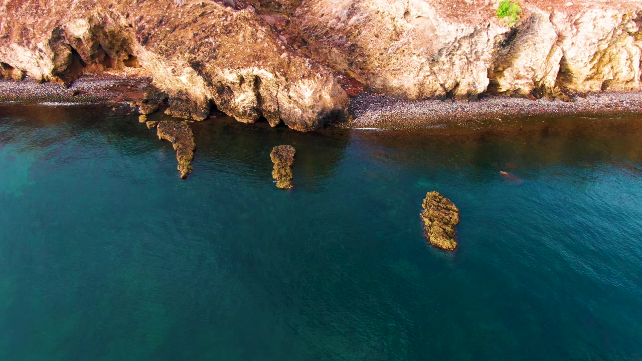 Aerial frame over Santa Rosalia showing the cliff edge across the top, two protruding rock formations near shore, blue-green water below, and no visible people.