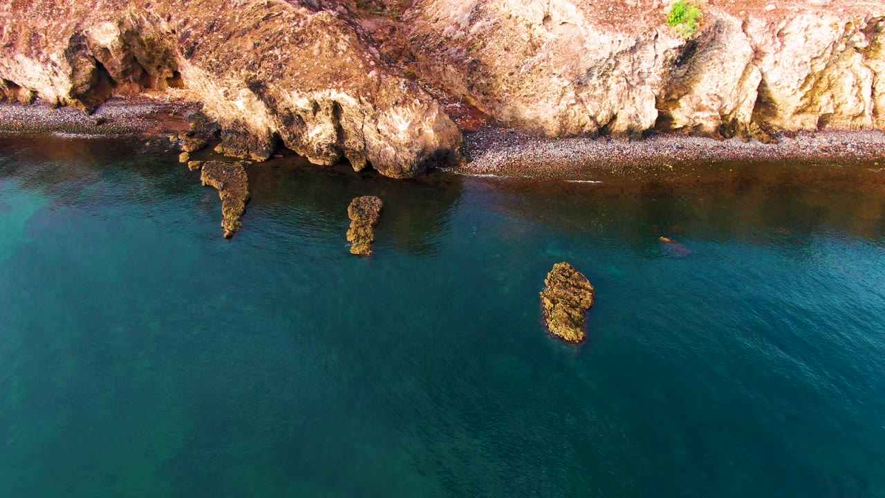 Overhead Santa Rosalia coastal view with rough tan rock along the upper frame, a thin dark shore line, clear water with subtle color shifts, and no visible people.