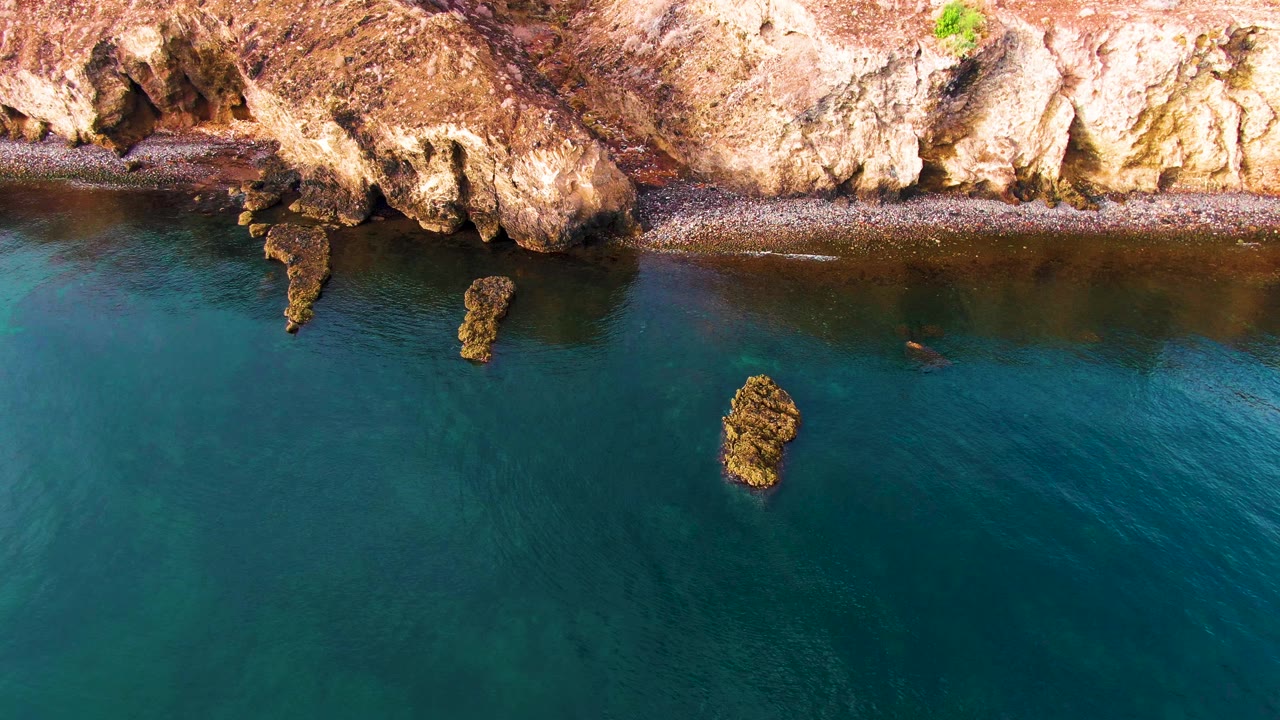 Aerial view of the Santa Rosalia coast with a pebbly beach strip under pale rock, darker teal water filling the lower frame, offshore rocks, and no visible people.