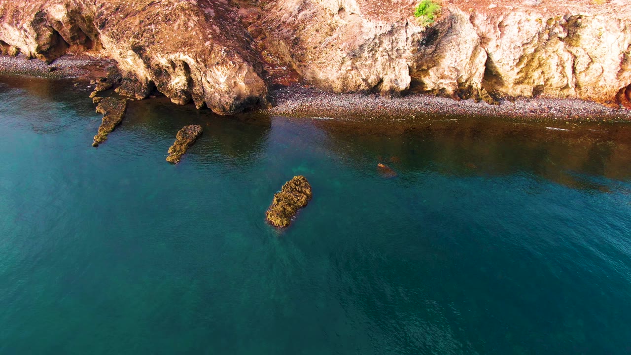 Aerial view of rocky Santa Rosalia coastline with tan cliffs, a narrow pebble shore, clear blue water, offshore rocks, and no visible people.