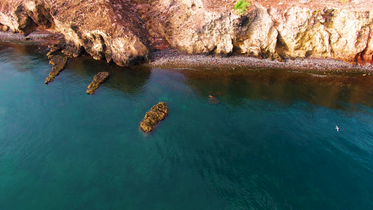 Overhead frame of the Santa Rosalia shore with reddish tan cliffs across the top, teal water below, several dark rock outcrops, and no visible people.