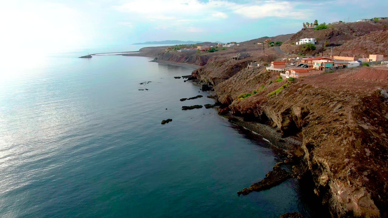 Aerial frame from Santa Rosalia, Baja California Sur showing calm blue-green water beside rocky cliffs, red-roofed homes on the ridge, and no visible people.