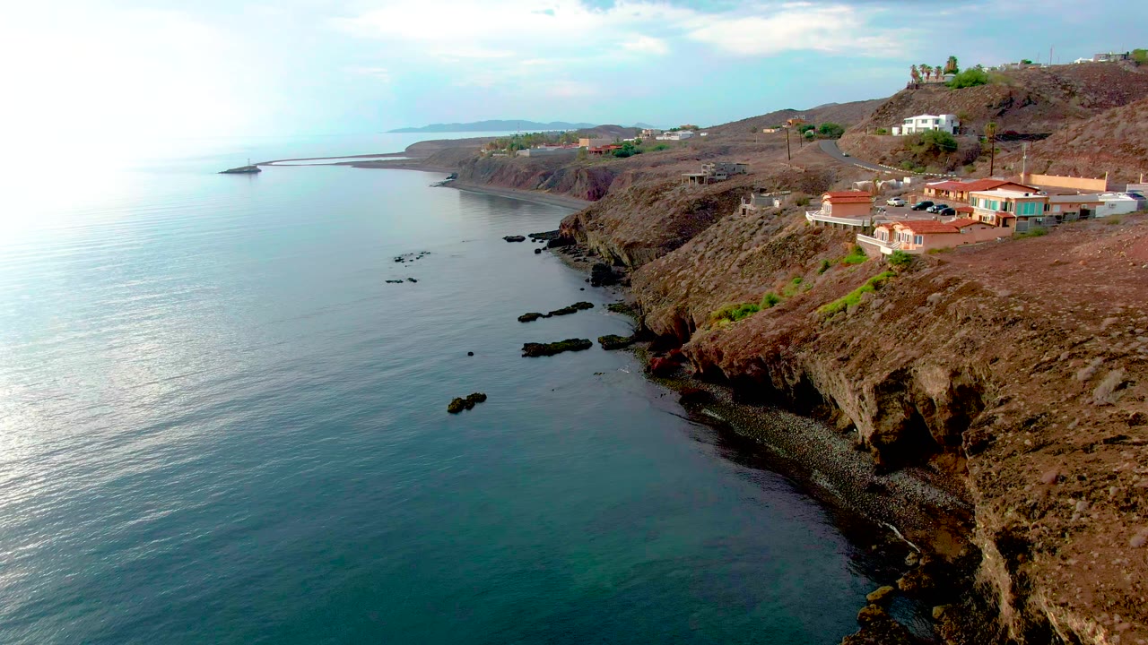 Aerial view in Santa Rosalia with more open water in the foreground, scattered black rocks near shore, dry brown cliffs, ridge-top houses, and no visible people.
