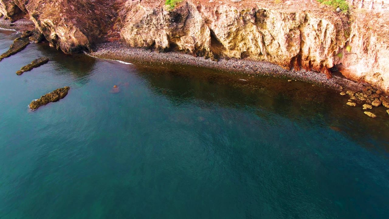 Aerial frame over Santa Rosalia shoreline, with dark nearshore water, a pebbly edge below the cliff, an offshore rock, and no visible people.