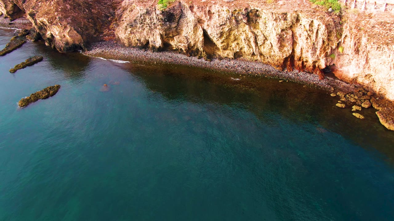 Aerial view of Santa Rosalia coast with blue-green water below a sunlit rock wall, small rocks near the right shoreline, and no visible people.