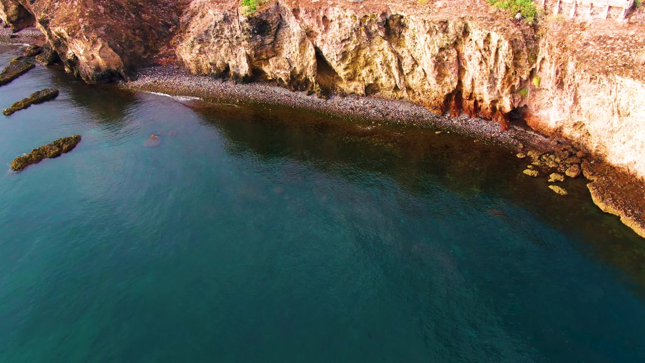 Aerial frame of Santa Rosalia rocky coast, where calm blue-green water curves below the cliff and small yellow-brown rocks cluster near the right edge, with no visible people.