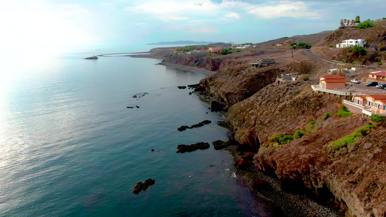 Aerial frame over Santa Rosalia, Baja California Sur, with calm blue water on the left, jagged brown cliffs on the right, hillside homes and a road above the shore, and no visible people.