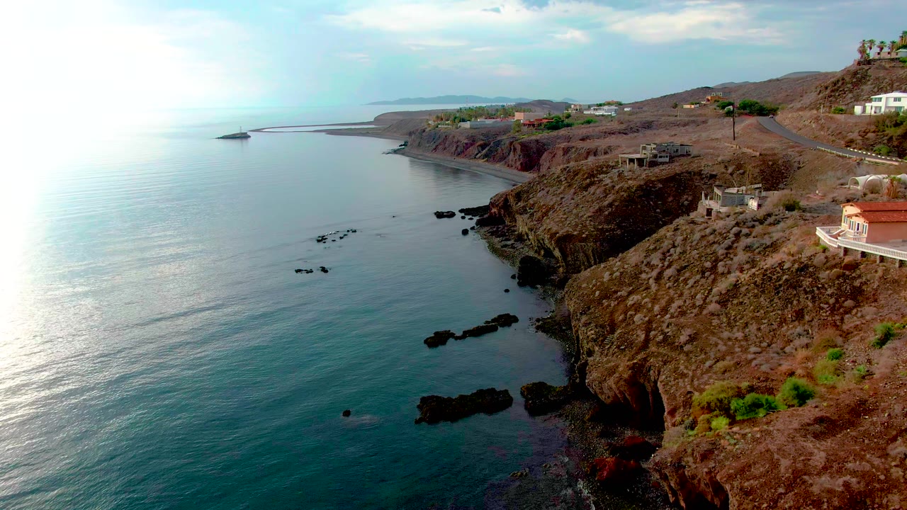 Aerial frame in Santa Rosalia showing calm blue-green sea, dark rocks near shore, brown cliff faces, a road along the hillside, several small buildings, and no visible people.