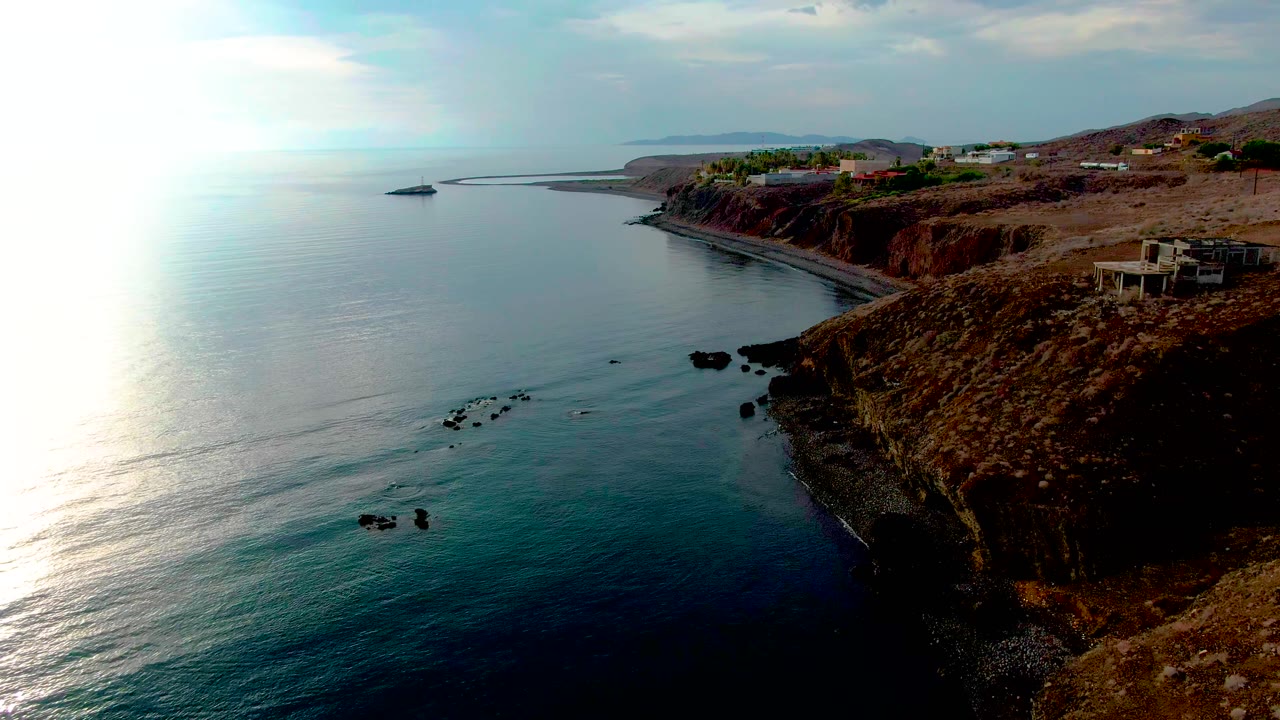 Aerial view of the Santa Rosalia coastline with blue ocean at left, reddish cliffs at right, cliffside buildings in the distance, sun reflection on the water, and no visible people.