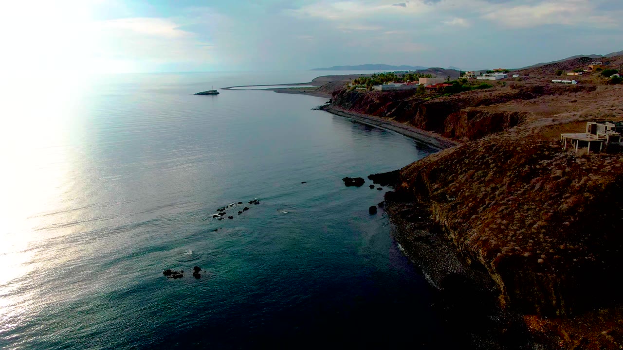 Santa Rosalia coastal frame with calm blue-green water, scattered dark rocks below the cliff, a bright reflection band on the sea, cliffside buildings, and no visible people.