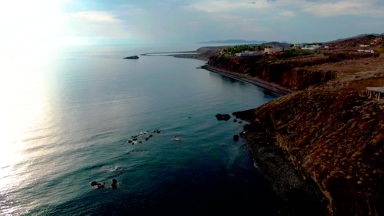 Aerial Santa Rosalia view with the cliff pushed farther right, broad sunlit sea across the frame, small rock clusters near shore, distant coastal structures, and no visible people.