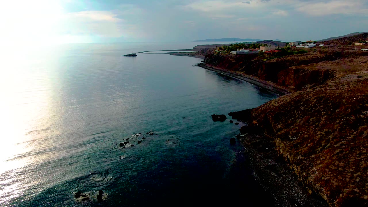 Coastal water with bright reflection, dark rocks, cliffs, and buildings above shore.