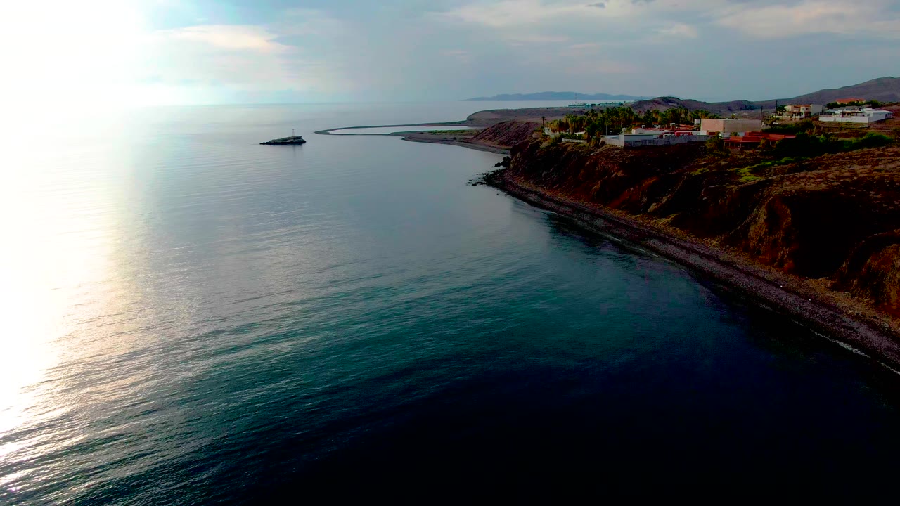 Opening aerial frame over the Santa Rosalia coastline, with bright glare on the ocean, a rocky cliff at right, hilltop buildings, distant land, and no visible people.