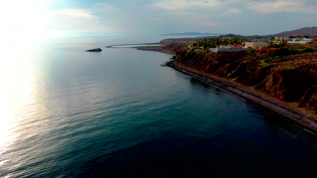 Early aerial view along Santa Rosalia's coast, showing calm blue water in the foreground, a diagonal pebbly shore, white blufftop buildings, and no visible people.