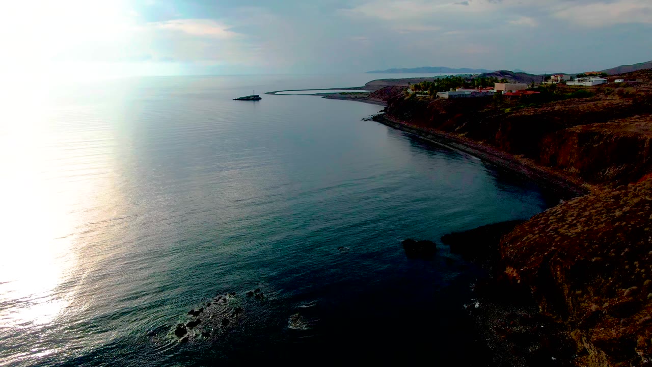 Wide aerial view in Santa Rosalia, Baja California Sur, with bright sun glare on the ocean at left, a rocky cliff at right, shoreline buildings above the water, and no visible people.