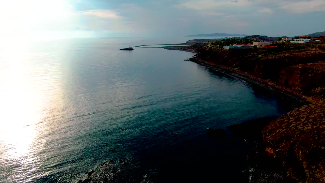 Blue ocean curves along a rocky cliff with a small offshore structure or boat near the coast.