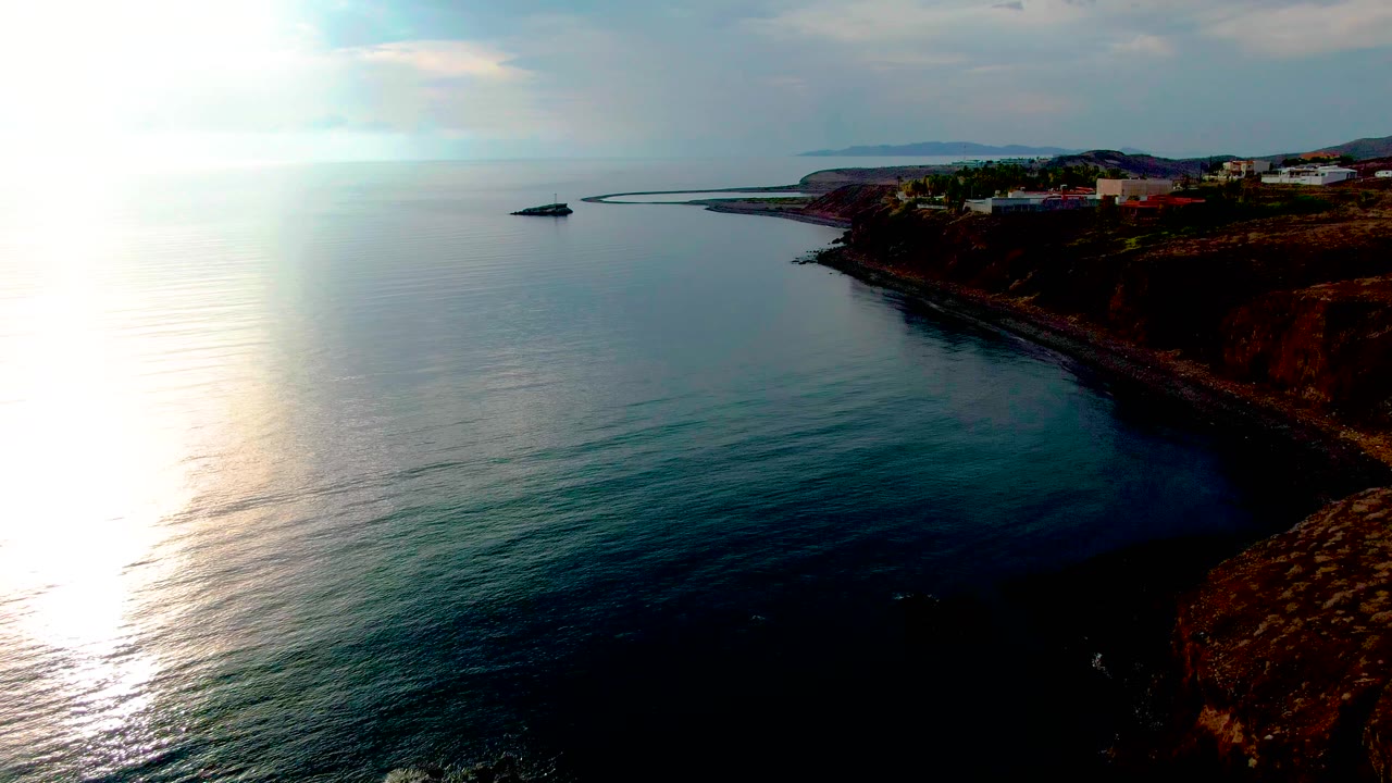 Aerial view over Santa Rosalia coast with the sunlit water dominating the left frame, dark calm water near the cliff, buildings on the upper right bluff, and no visible people.