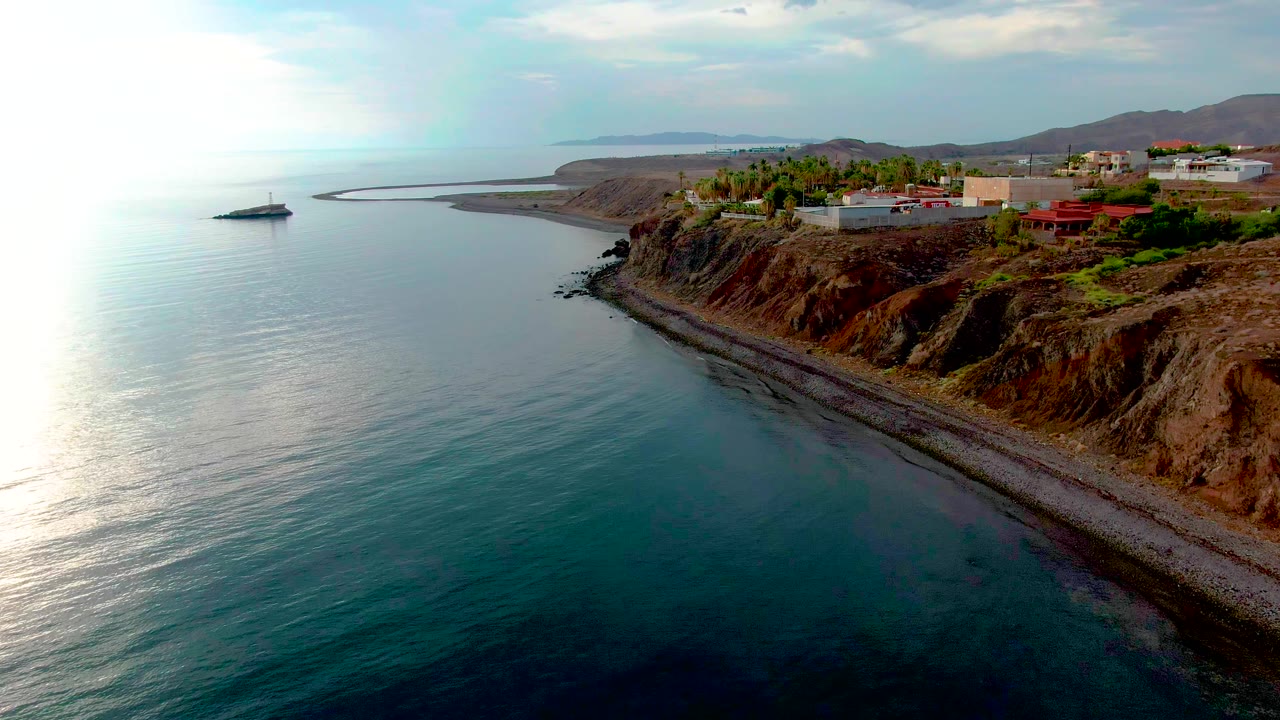Aerial frame over Santa Rosalia, Baja California Sur, showing calm blue-green ocean, a narrow pebble shore, reddish rocky cliff, blufftop buildings, a distant sailboat, and no visible people.