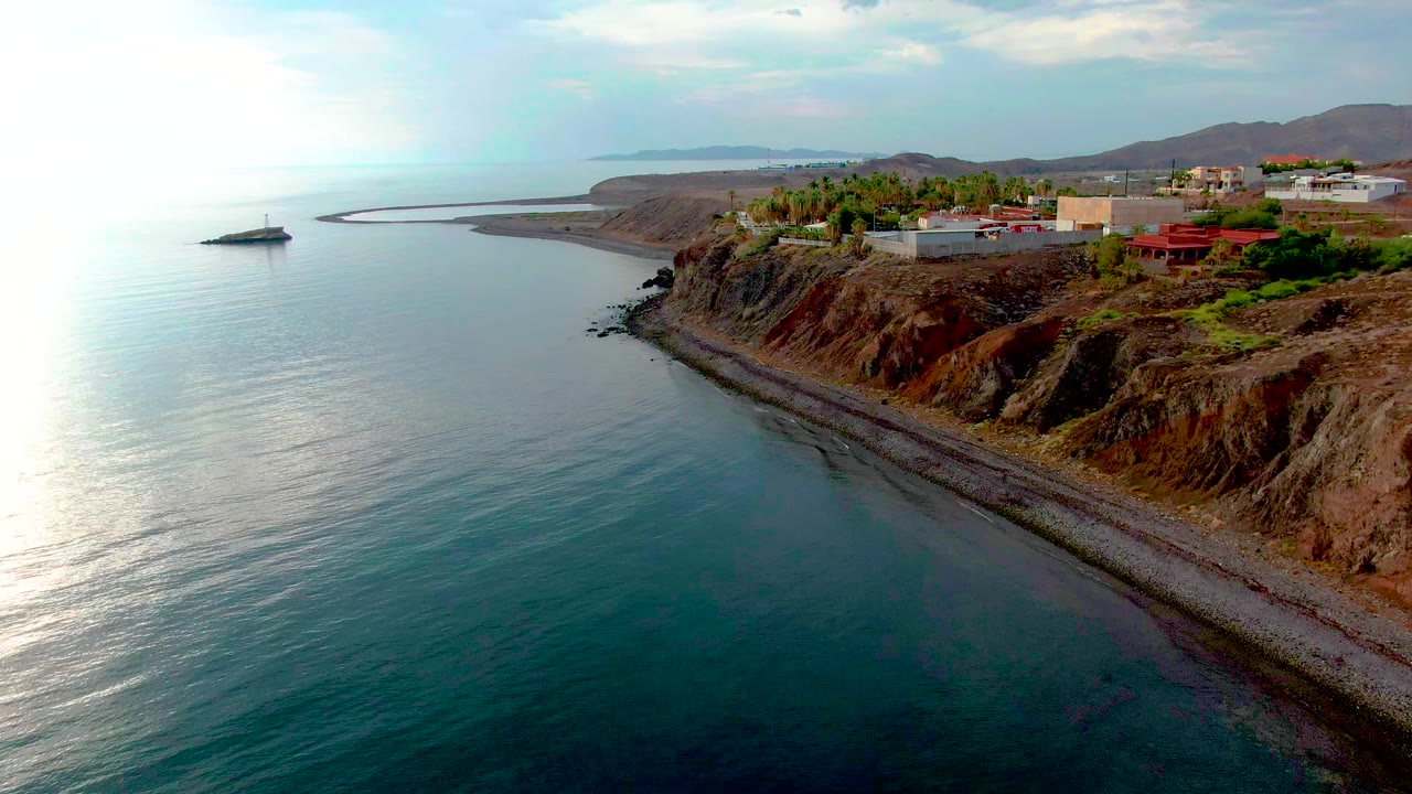 Aerial view in Santa Rosalia with the sailboat left of frame, bright reflection across the calm water, a curving pebble beach below the cliff, buildings on the bluff, and no visible people.