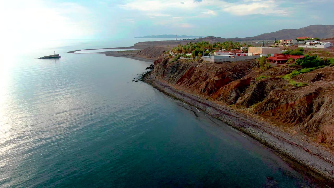 Aerial frame over the Santa Rosalia coast with smooth ocean water in the foreground, a narrow stony shore at the cliff base, blufftop buildings, distant landforms, and no visible people.