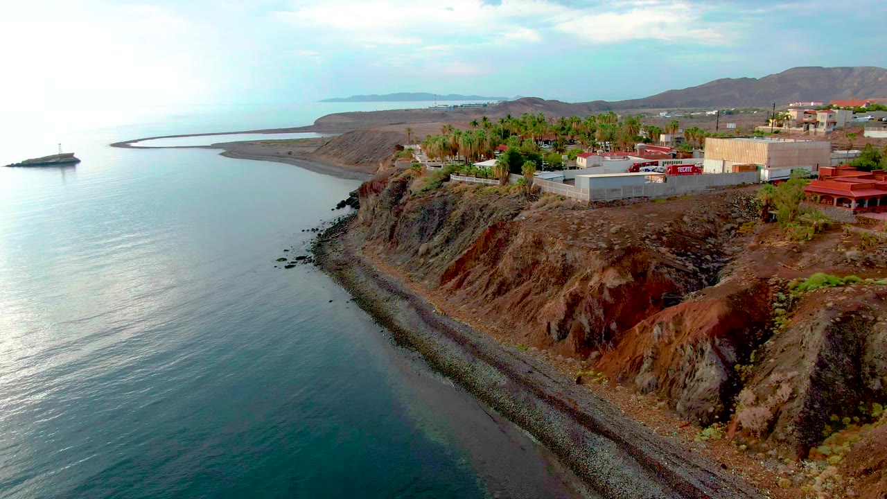Aerial frame over Santa Rosalia, Baja California Sur, showing calm blue-green water, a pebbly shore, red-brown cliff faces, bluff-top buildings, palm trees, distant hills, and no visible people.
