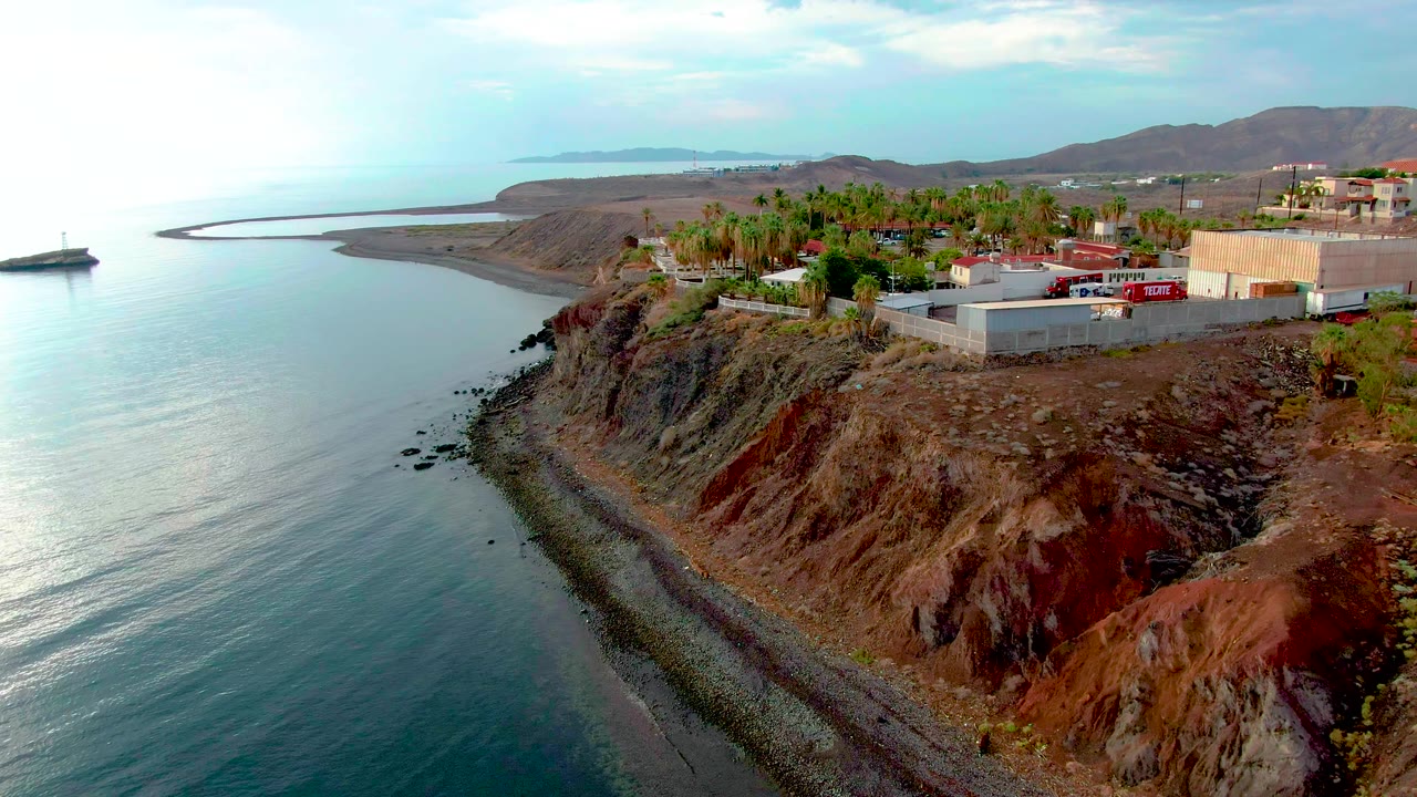 Aerial frame from Santa Rosalia showing eroded red and gray cliff layers above a narrow rocky beach, calm water to the left, buildings and palm trees on top, and no visible people.