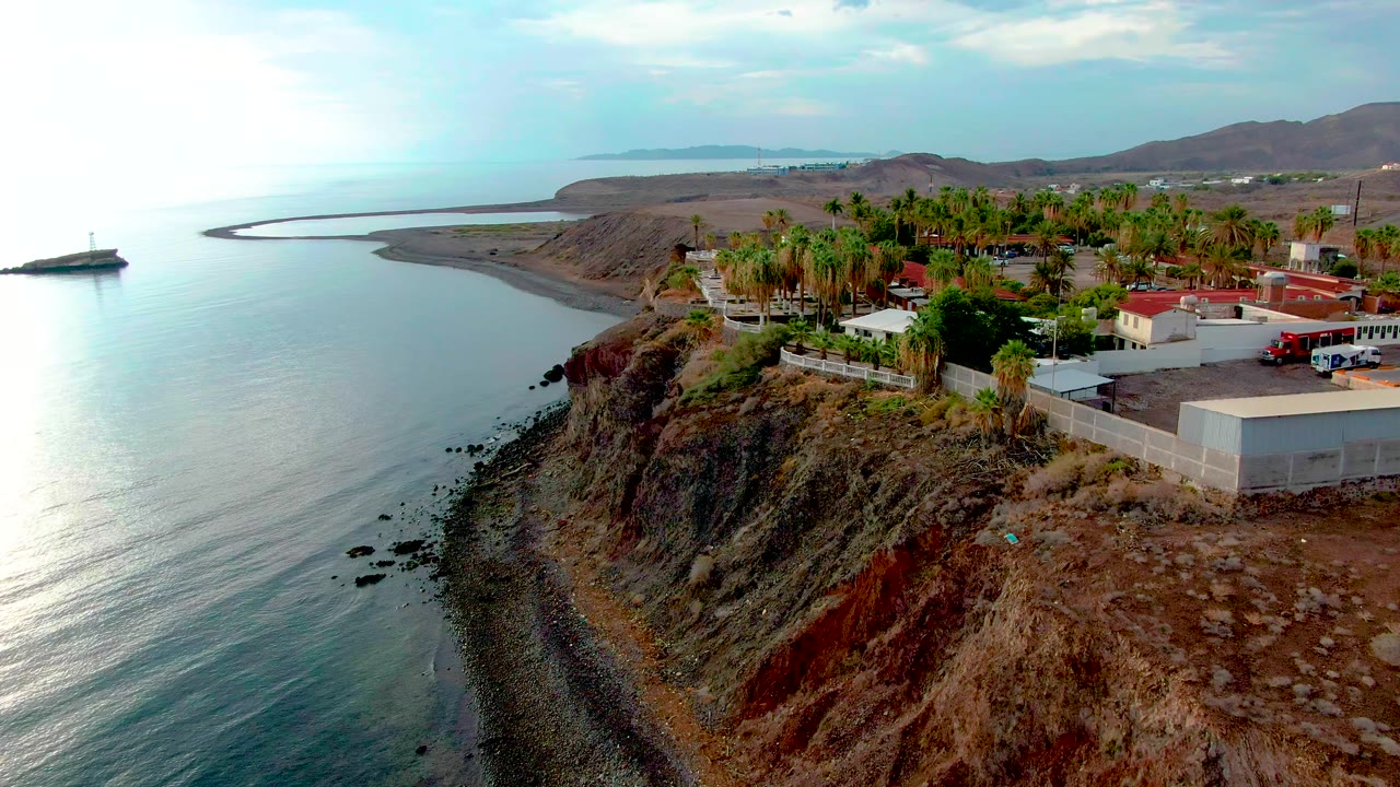Opening aerial frame over Santa Rosalia, Baja California Sur, with calm ocean at left, a rocky cliff below, palm trees and low shore buildings at right, and no visible people.