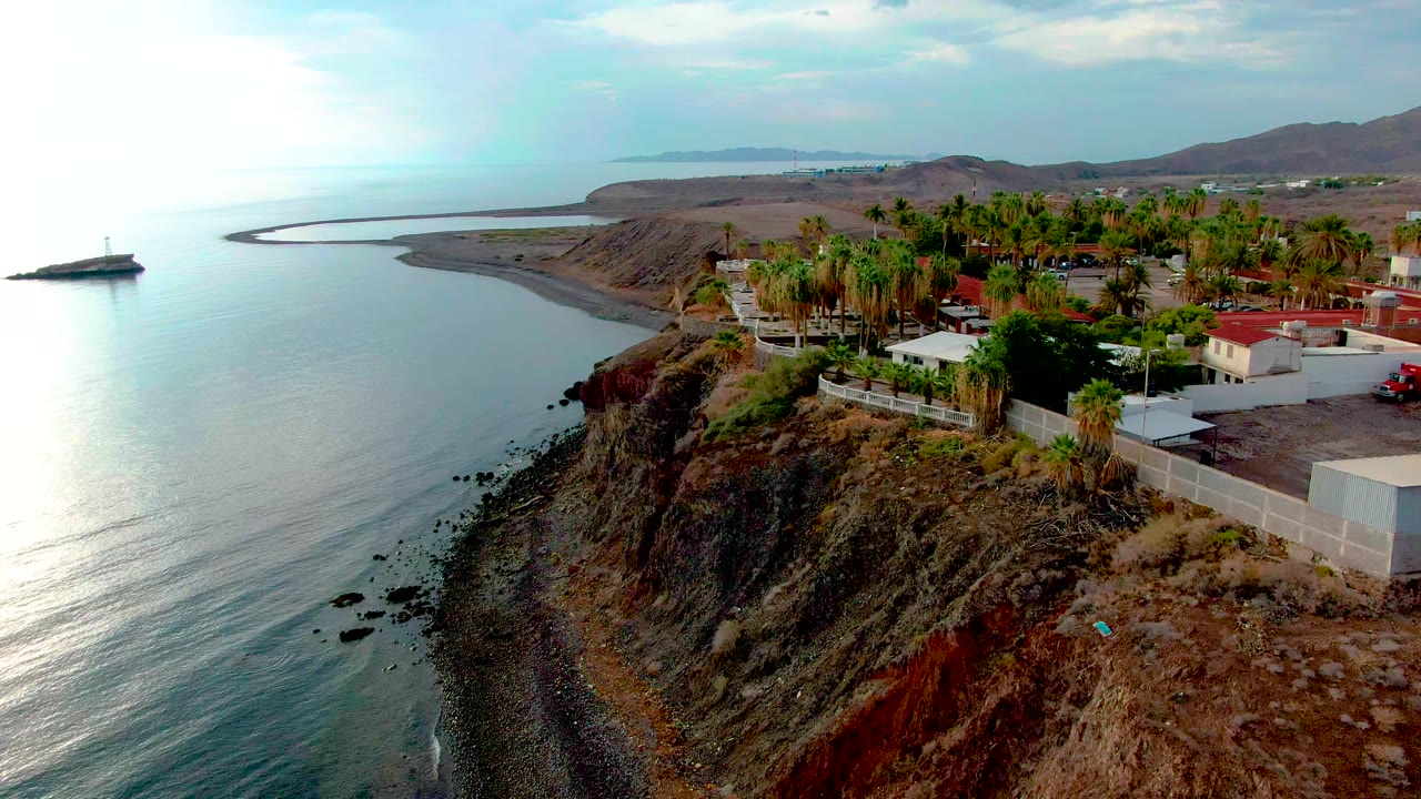 Aerial frame in Santa Rosalia showing the ocean curving along the rocky shore, with palms and low buildings on the cliff top and no visible people.