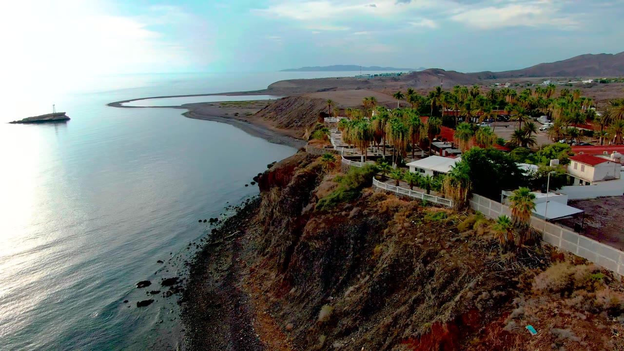 Drone-style view of Santa Rosalia coastline with bright water reflection at left, eroded cliff in the center, palms and buildings at right, and no visible people.