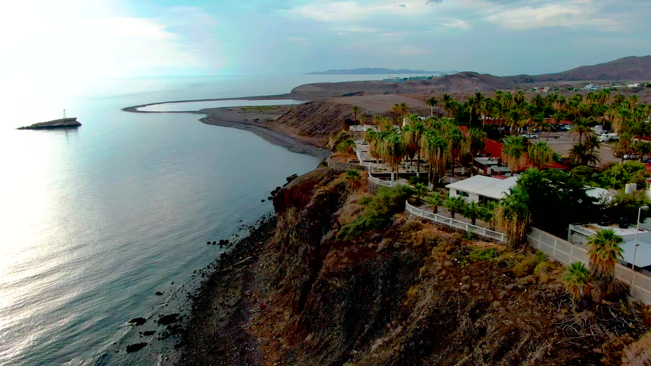 Wide aerial coastline with cliff, ocean, palm trees, and a small offshore vessel.
