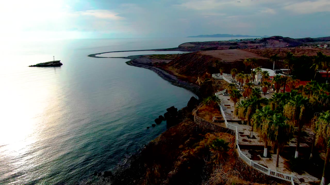 Aerial frame of Santa Rosalia coastline with bright sun reflection on the ocean, a small offshore marker, a cliffside path lined with palms, and no visible people.