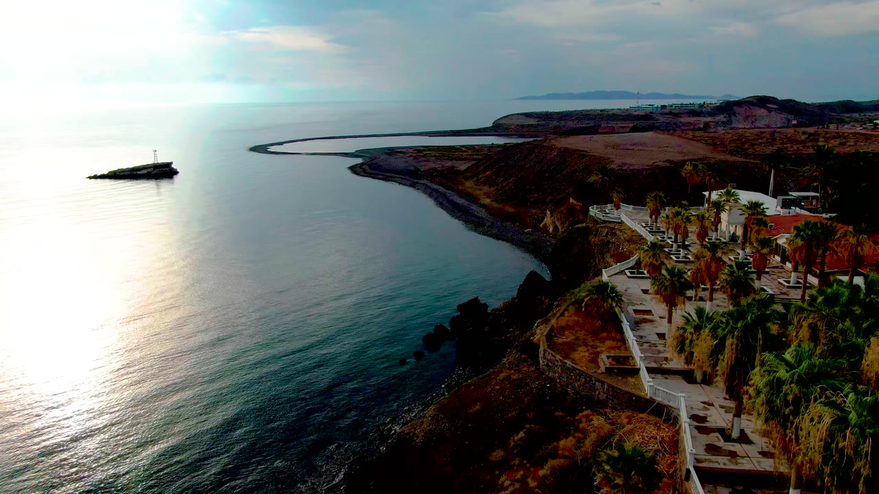 Aerial view in Santa Rosalia with the palm-lined cliff path still prominent at right, the ocean spread across the left, a rocky shore below, and no visible people.