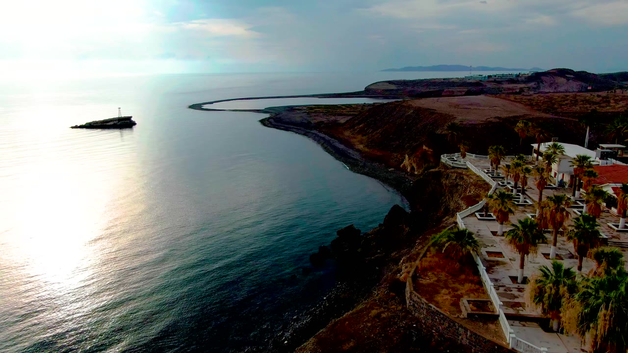 Santa Rosalia aerial frame showing teal water beside a dark rocky shoreline, a white path with palms near the cliff edge, distant land across the horizon, and no visible people.