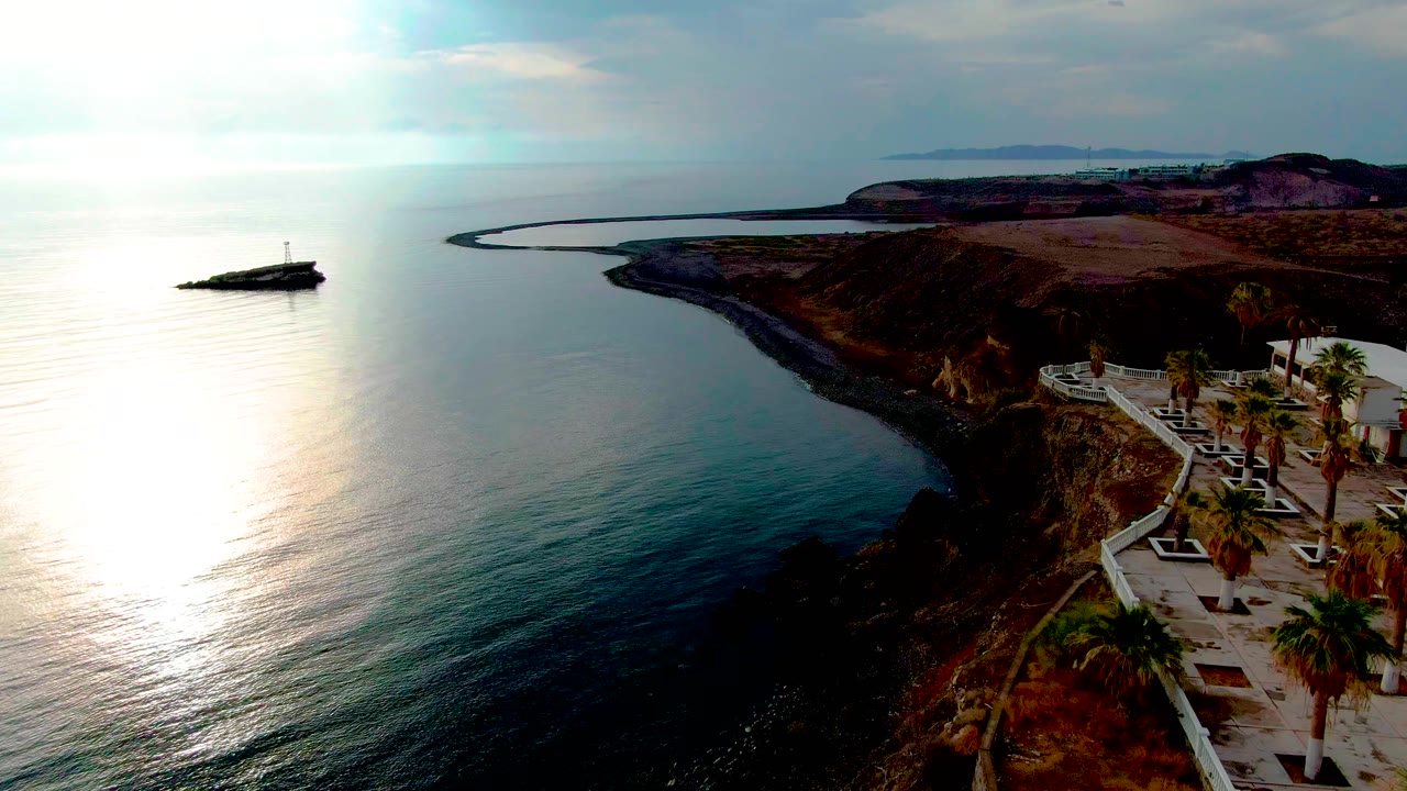 Aerial frame from Santa Rosalia with intense sunlight reflected on the water, a small offshore beacon at left, the curved rocky coast through center, and no visible people.