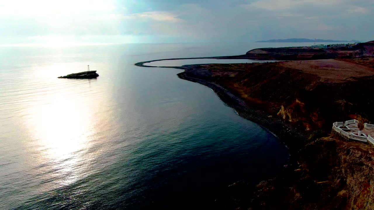 Aerial view of the Santa Rosalia coastline with bright sun reflection on the ocean, a dark rocky shore at right, distant land on the horizon, and no visible people.