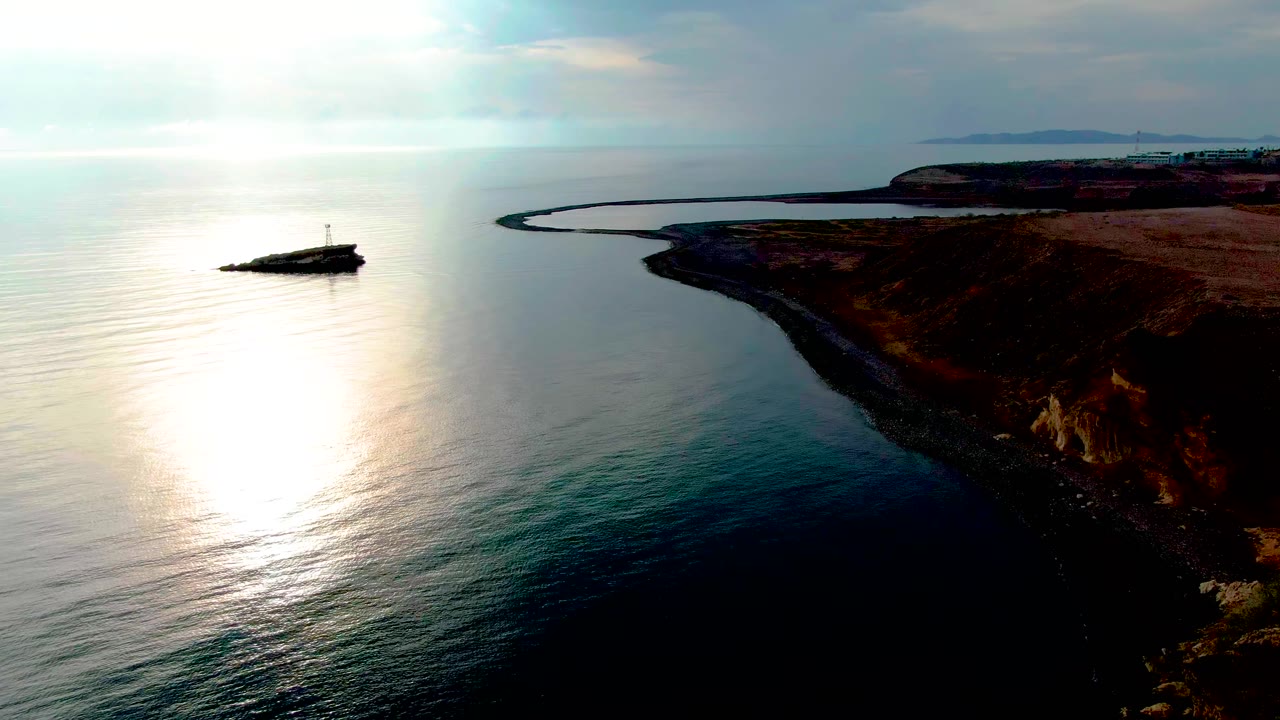 Aerial view of Santa Rosalia's rocky coast with a small offshore marker or breakwater left of center, bright reflected sunlight on the water, and no visible people.