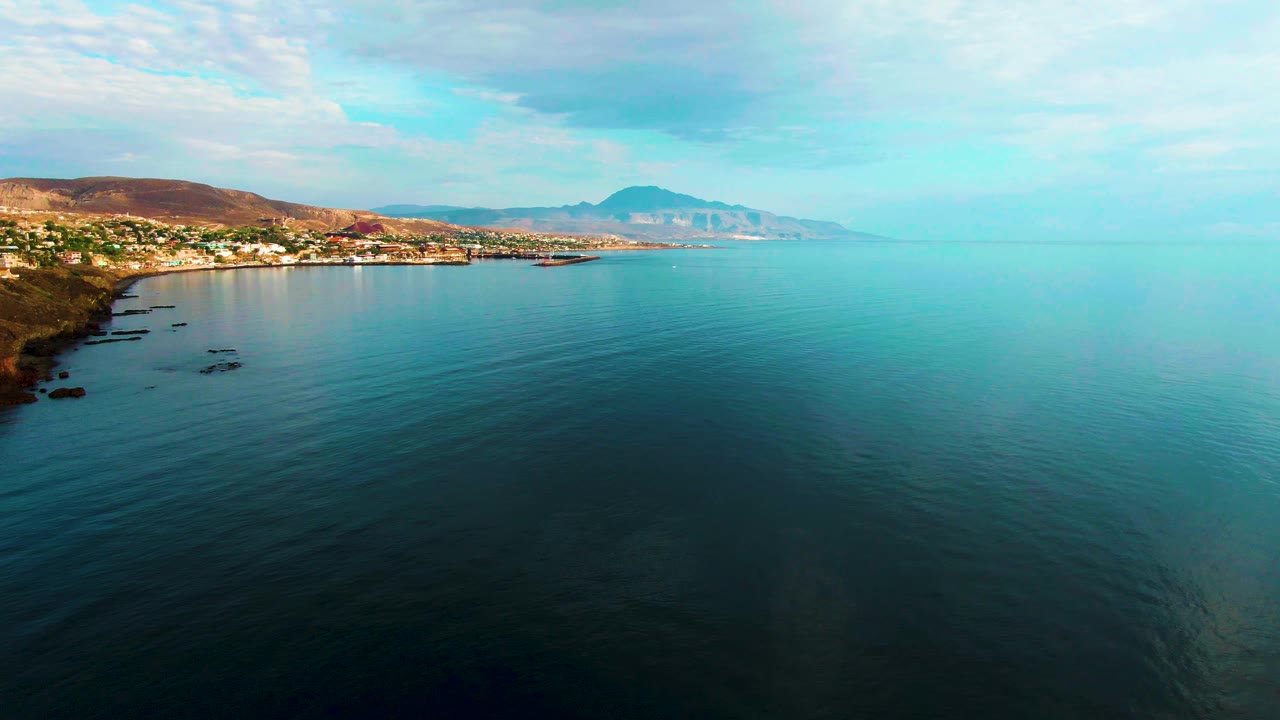 Opening frame of Santa Rosalia, Baja California Sur with calm blue water in the foreground, rocky coast at left, hillside buildings in the distance, and no visible people.