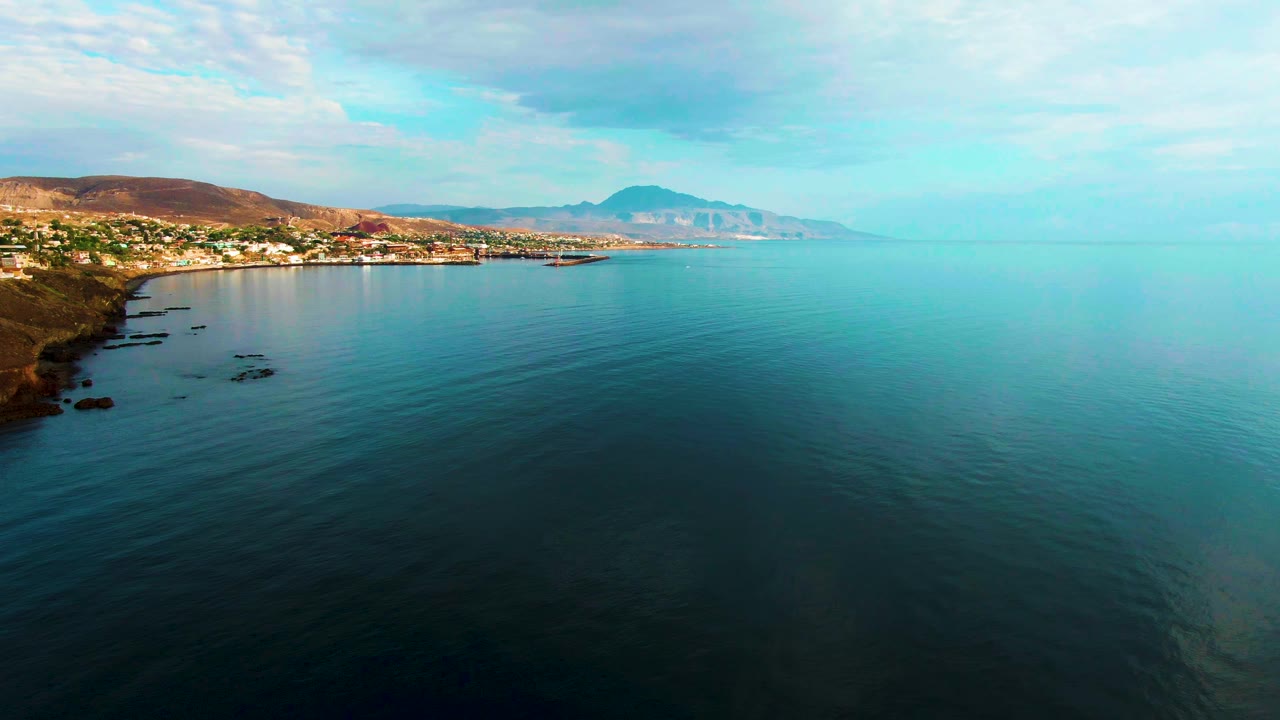 Wide frame over Santa Rosalia water with the shoreline and buildings held to the left, distant hills across the horizon, soft clouds overhead, and no visible people.