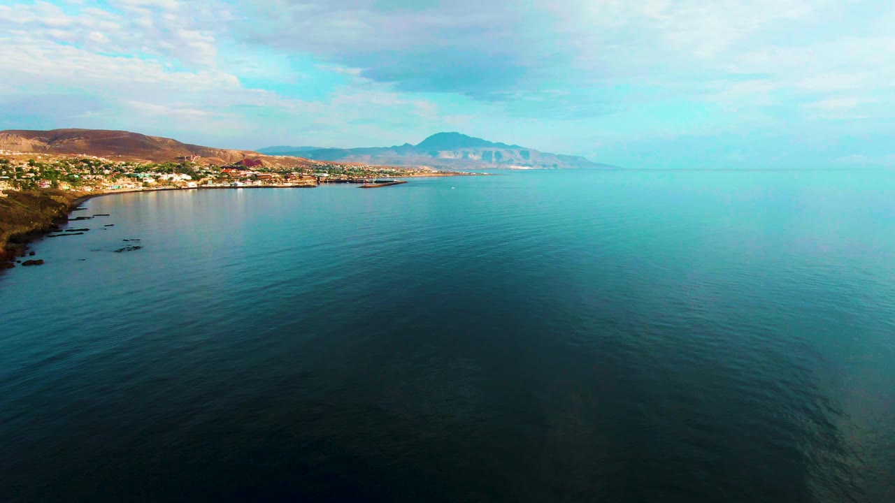 Aerial frame of Santa Rosalia coastline with smooth ocean ripples in the foreground, small shoreline buildings to the left, hazy mountains ahead, and no visible people.