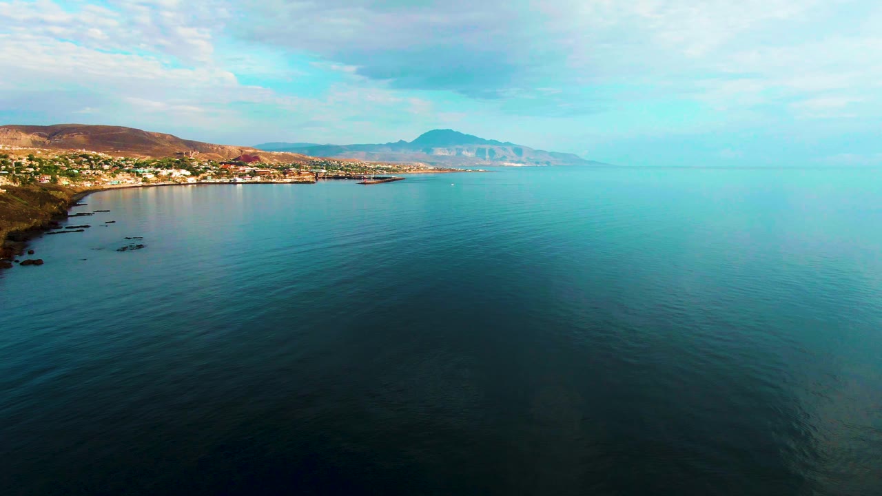 Aerial shoreline view with calm water, buildings, hills, and cloudy sky.