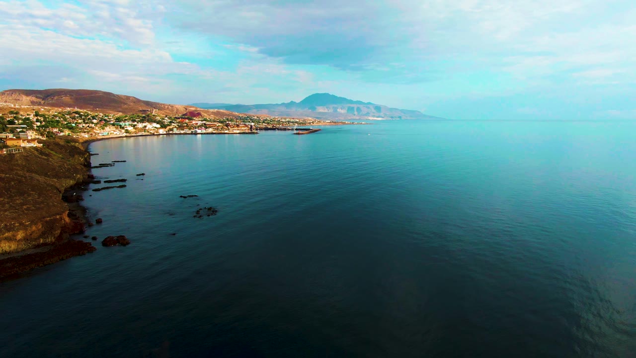 Aerial frame over Santa Rosalia with a rocky headland at left, small coastal buildings in the upper left, calm turquoise water occupying most of the view, hazy hills on the horizon, and no visible people.