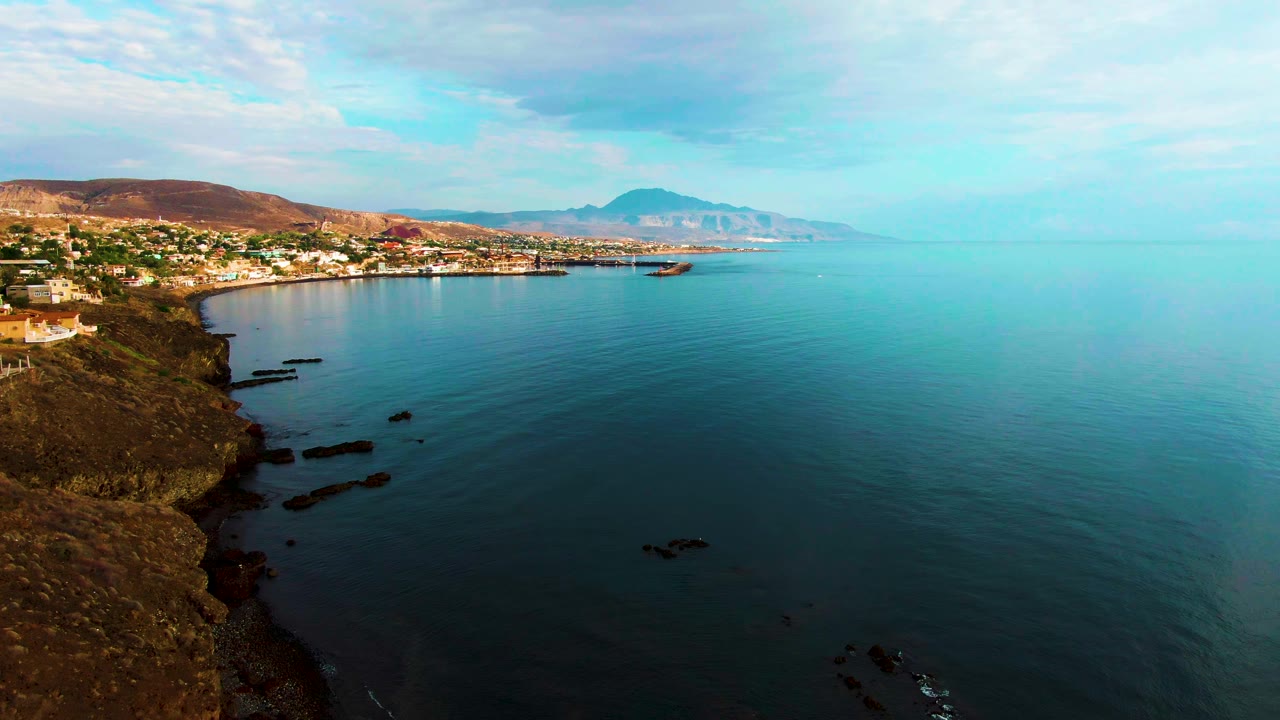 Opening frame of Santa Rosalia, Baja California Sur with rocky shoreline at left, calm blue-green coastal water, a small boat offshore, distant town and hills, and no visible people.