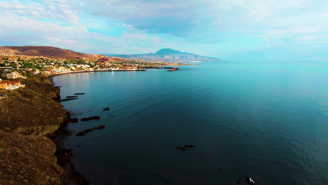Early frame of Santa Rosalia coastal water where the rocky left edge and town remain steady, the small boat sits farther out on the blue-green surface, and no visible people appear.