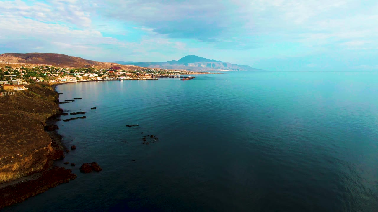 Aerial frame of Santa Rosalia, Baja California Sur, Mexico, with a rocky cliff on the left, calm blue ocean to the right, a small coastal town across the bay, distant hills, and no visible people.