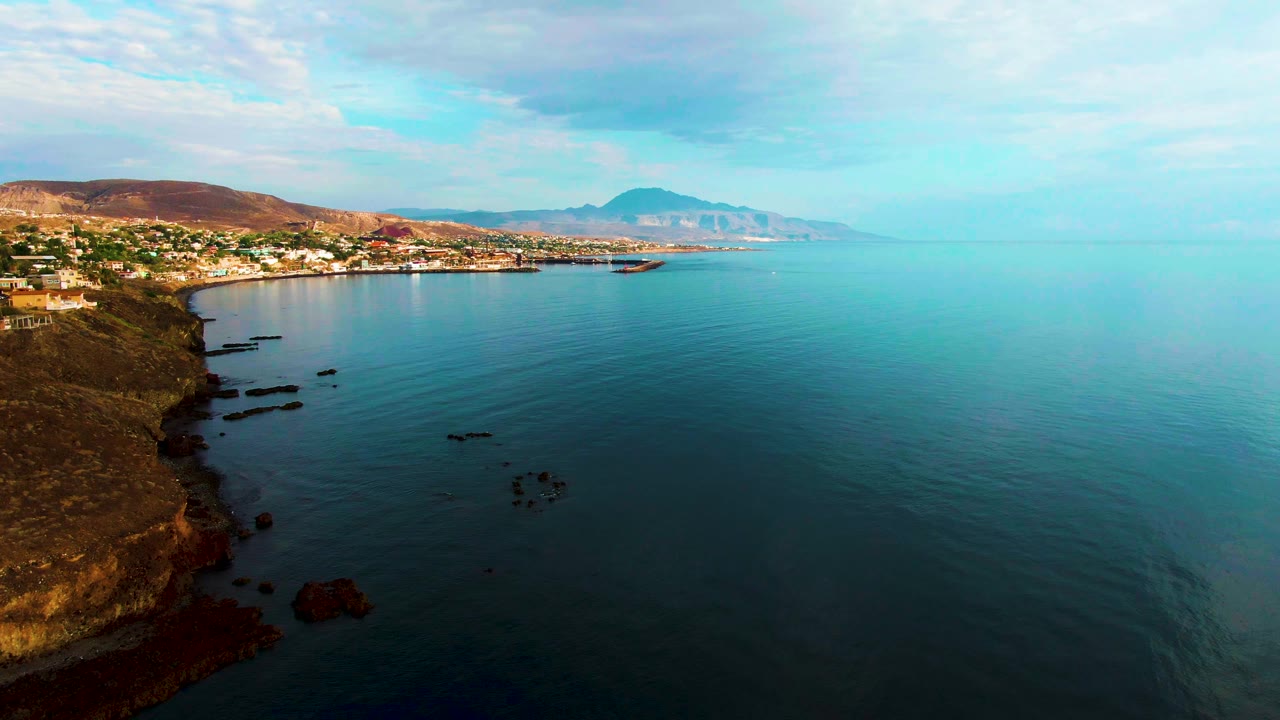 Santa Rosalia shoreline seen from above, with brown rock ledges at left, smooth blue water across the frame, white buildings along the bay, hazy hills in back, and no visible people.
