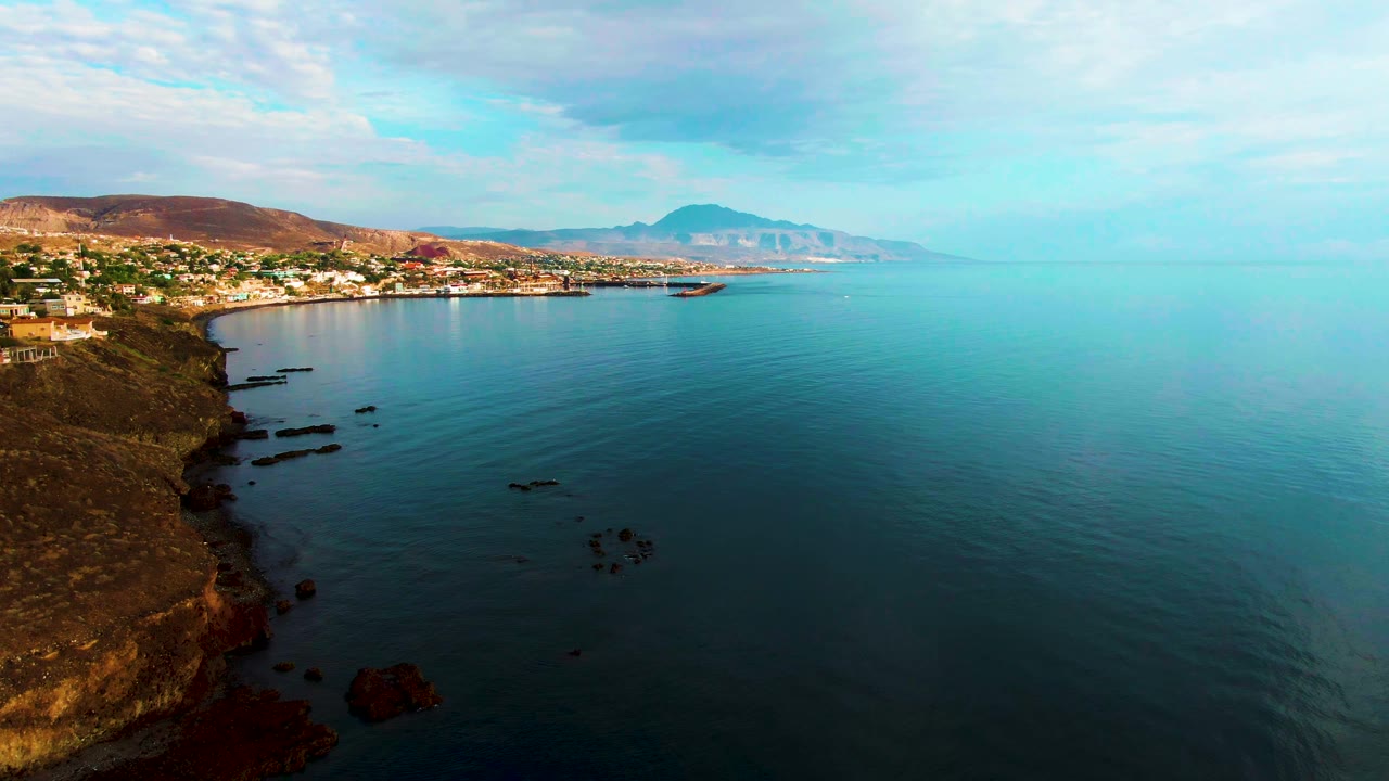 A high aerial view over Santa Rosalia with the cliff edge dropping into dark rocks, open turquoise and blue water, a line of town buildings, distant mountain shapes, and no visible people.