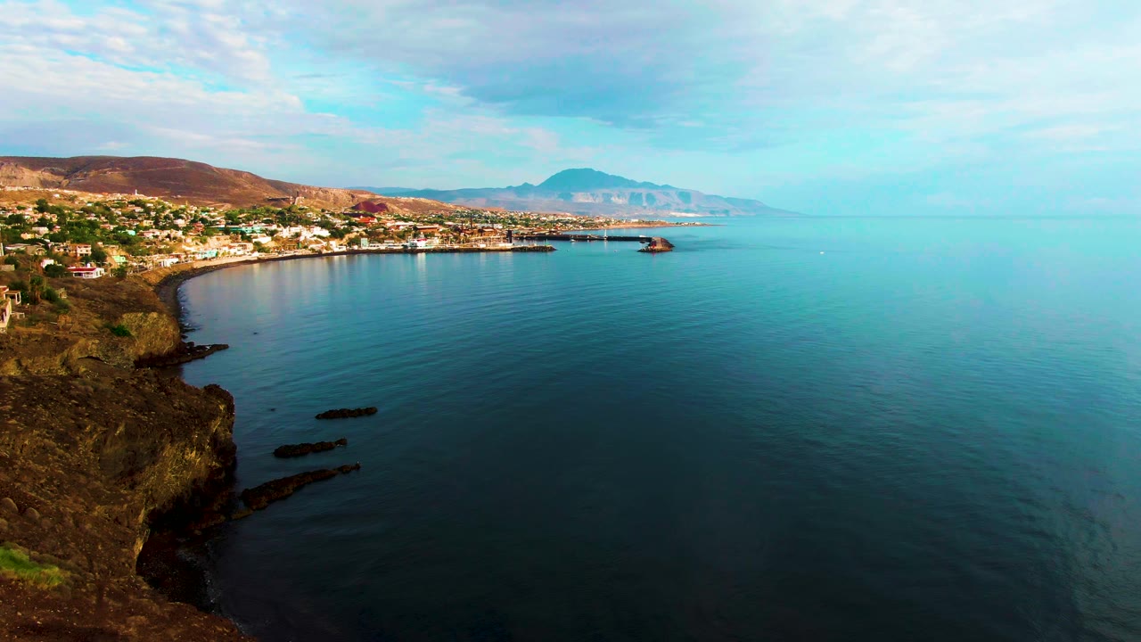 Opening aerial frame over Santa Rosalia, Baja California Sur, with a rocky cliff at left, calm blue ocean to the right, shoreline buildings across the bay, distant mountains, and no visible people.