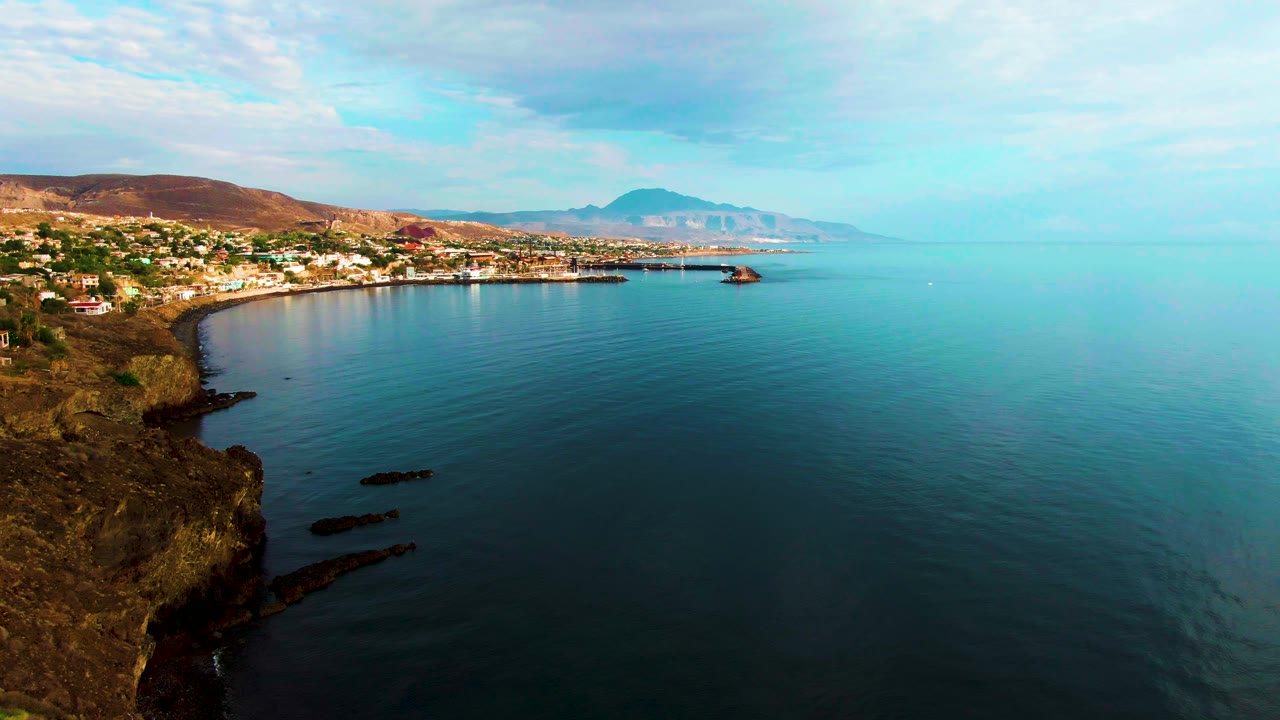 Aerial frame over Santa Rosalia showing the cliff edge lower left, dark nearshore rocks in the water, a curved line of coastal buildings, hazy mountains, and no visible people.