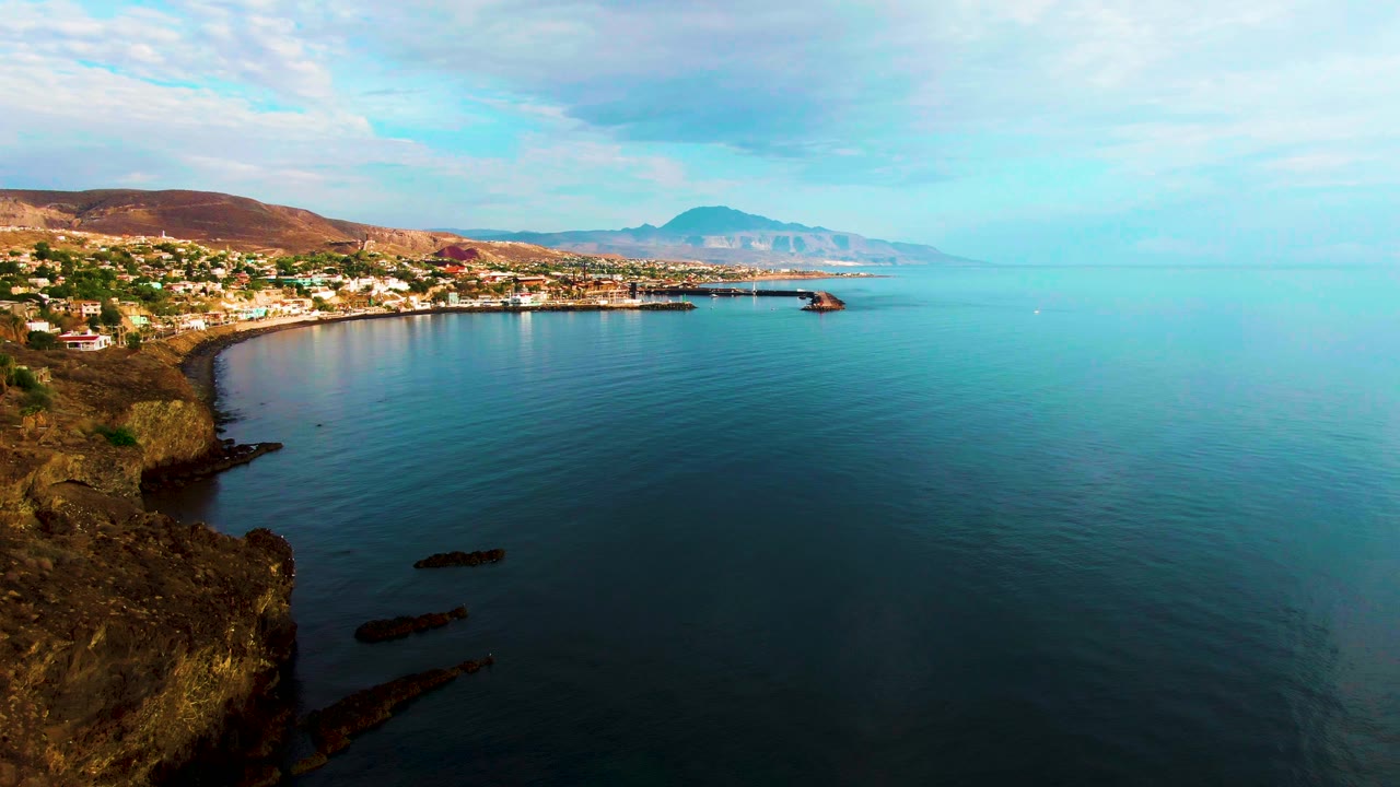Coastal cliff and bay with calm water, distant buildings, and mountains under clouds.